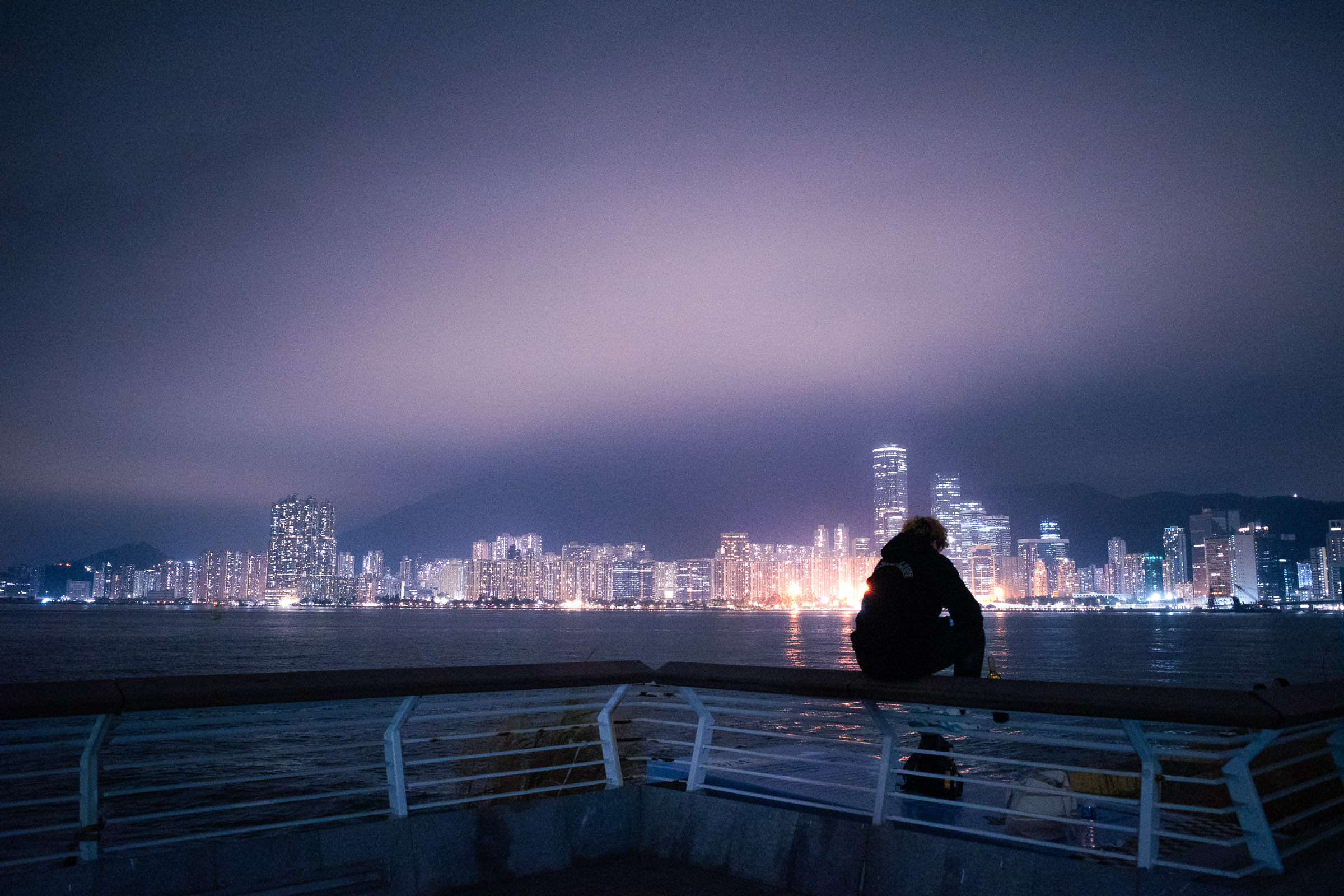 A man sits on some railings, looking out at the Hong Kong skyline at night. The City lights of Hong Kong are visible in the background